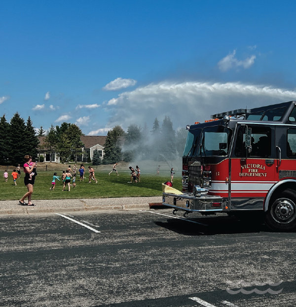 Fire truck sprays water on a field with people running through the water.
